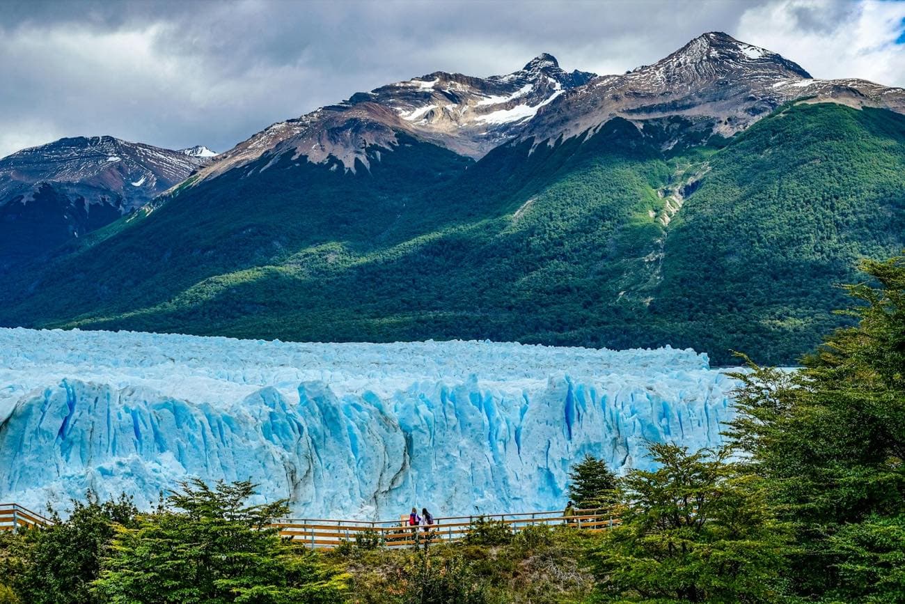 The blue face of the Perito Moreno glacier towering over the wooden boardwalks where two visitors walk, beneath the forested mountains of Los Glaciares park