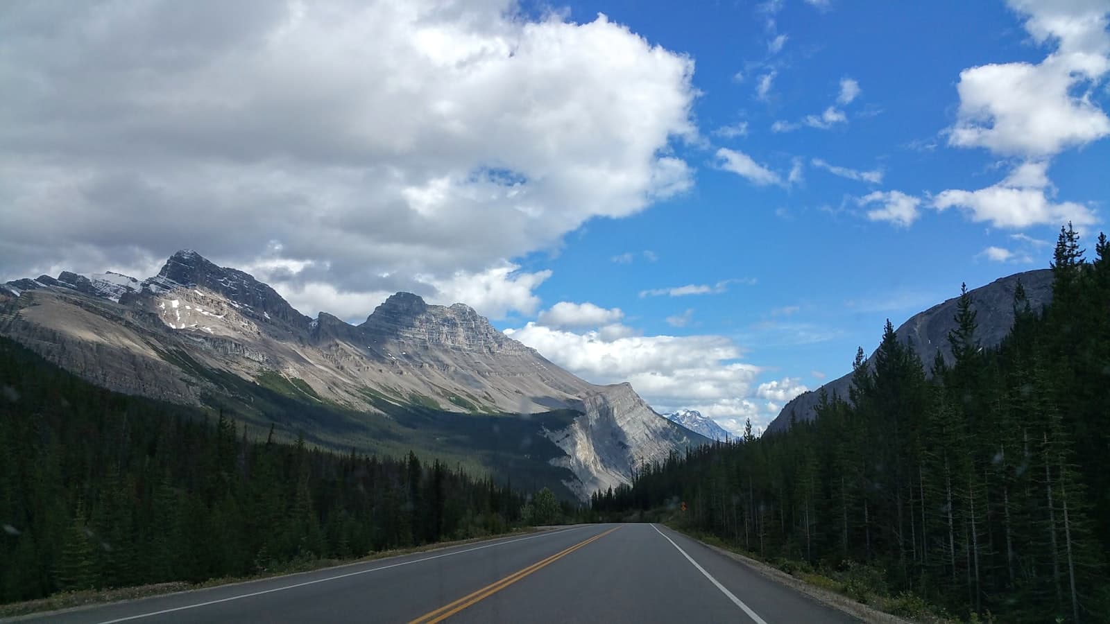 The Icefields Parkway running straight between spruce forests and Rockies walls under a cumulus sky