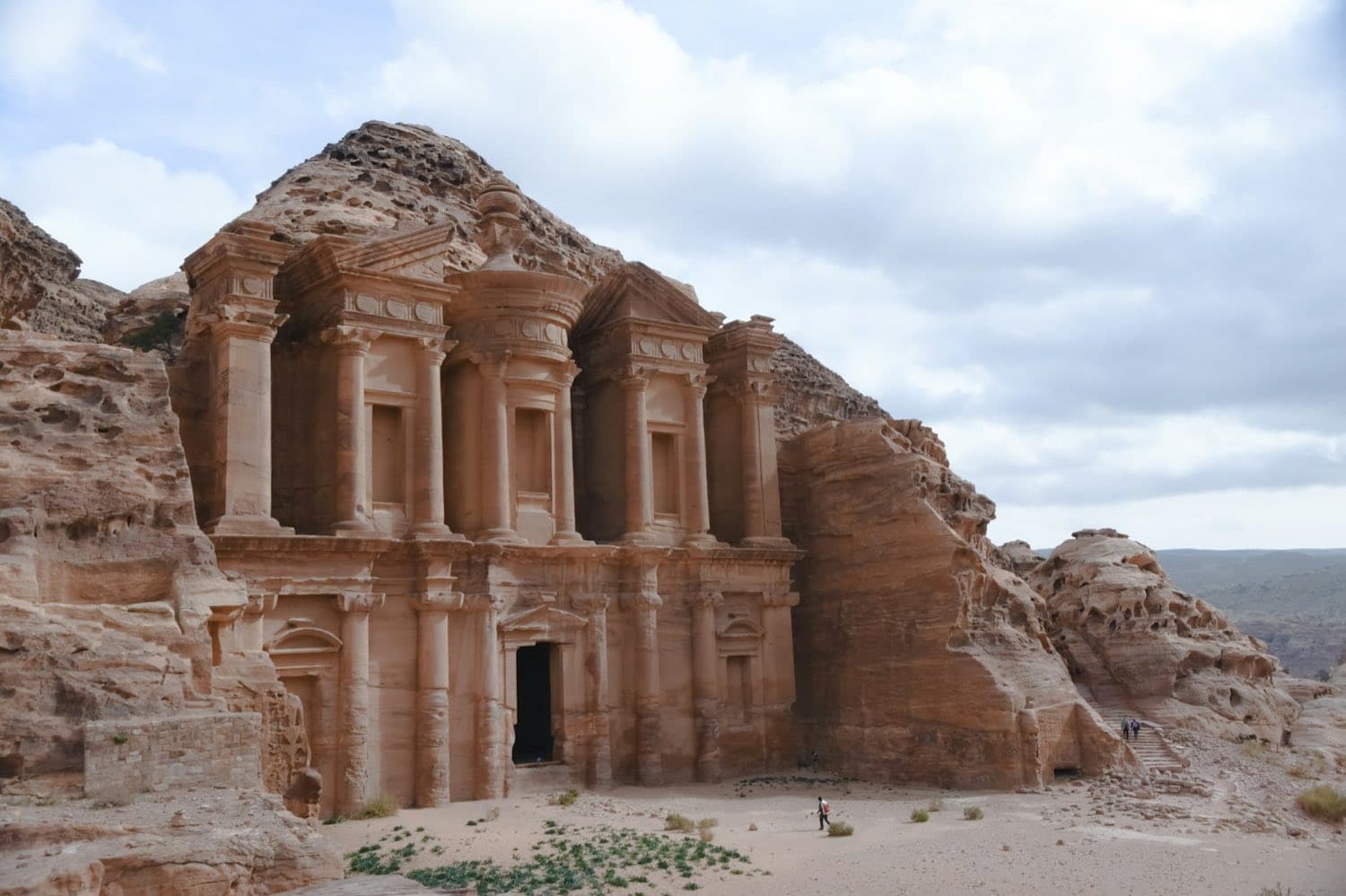 The colossal façade of the Monastery (Ad-Deir) carved into Petra's rose sandstone cliff, with tiny hikers at the foot of the monument