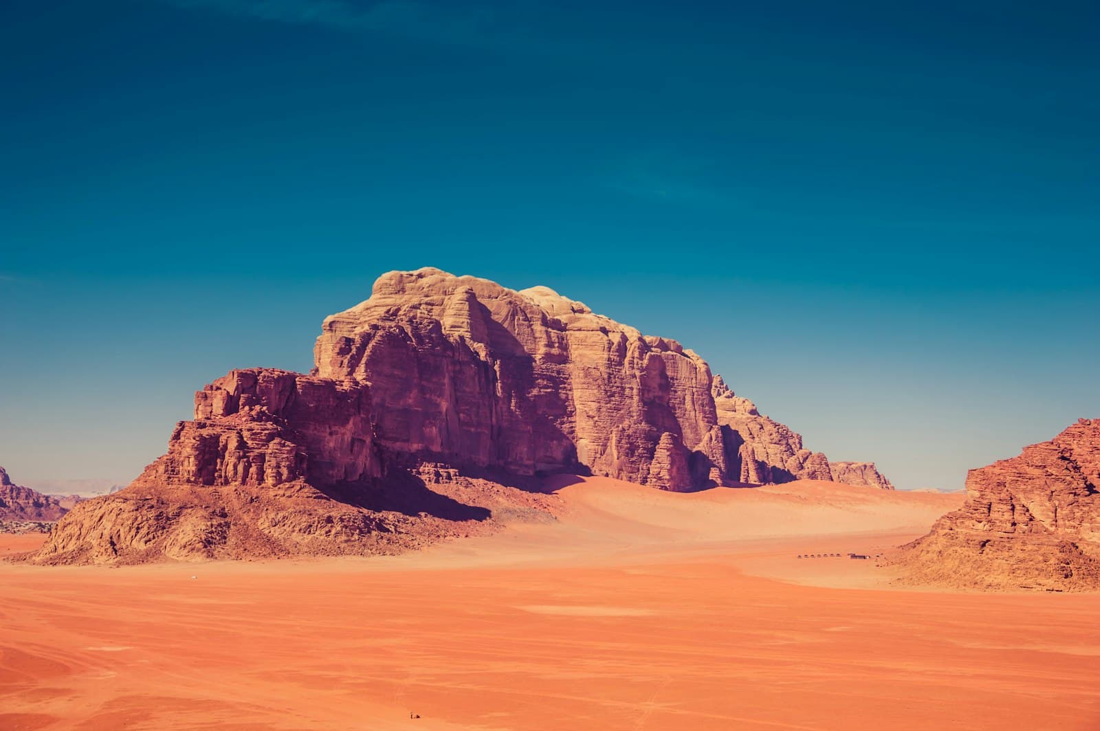 A sandstone mesa towering over the red sand of Wadi Rum under a deep blue sky, a tiny Bedouin camp at its base