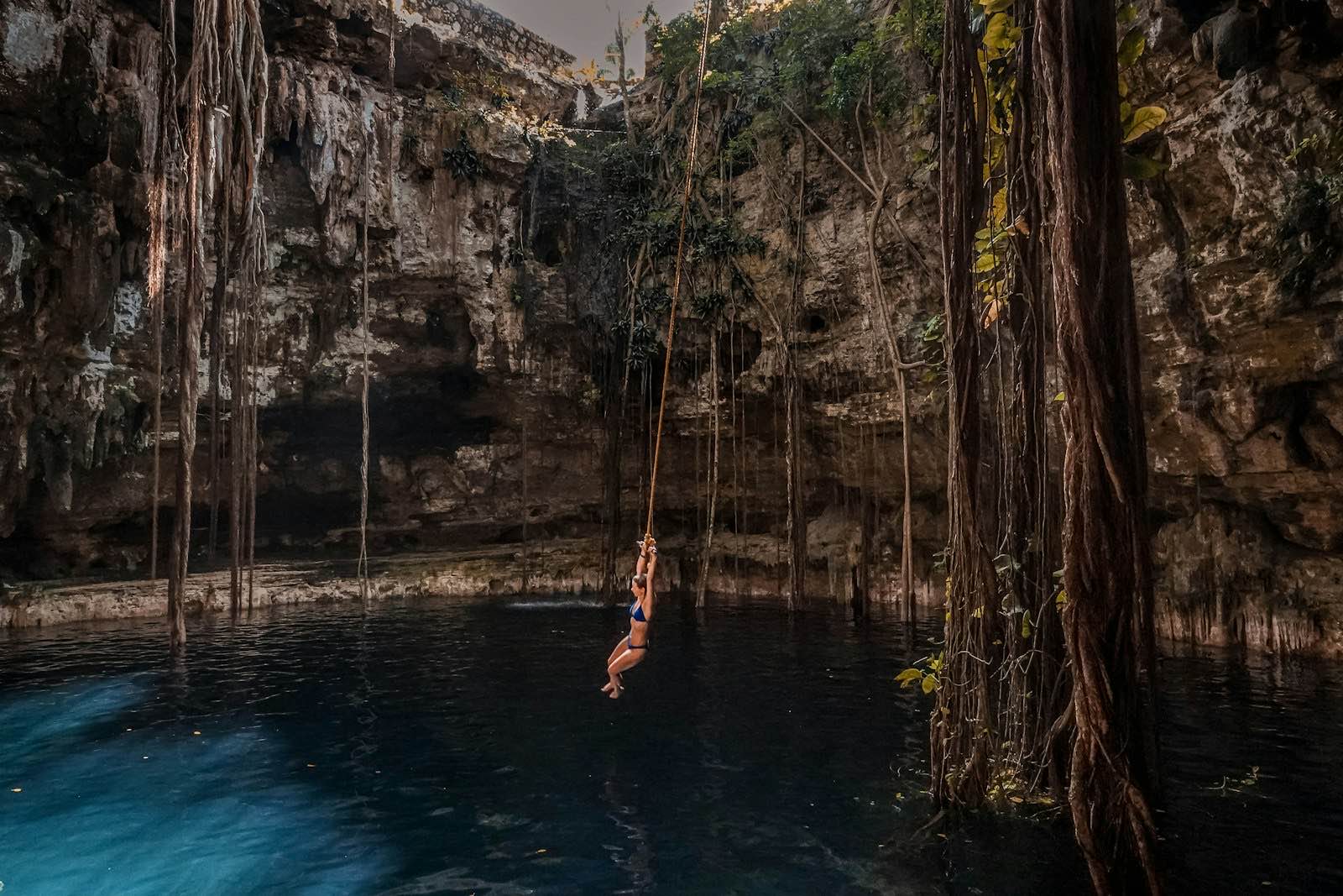 A swimmer hanging from the rope swing above the dark water of Cenote Oxman, beneath aerial roots falling from the collapsed cave roof