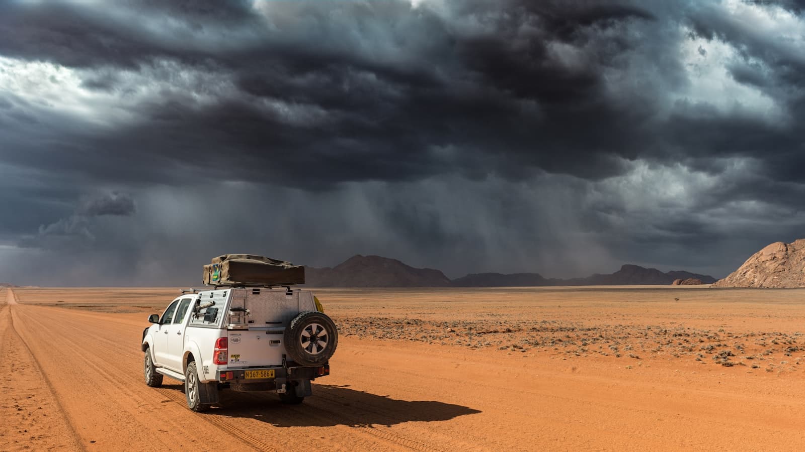 Un 4x4 équipé d'une tente de toit arrêté sur une piste de gravier rouge du Namib, sous un ciel d'orage spectaculaire
